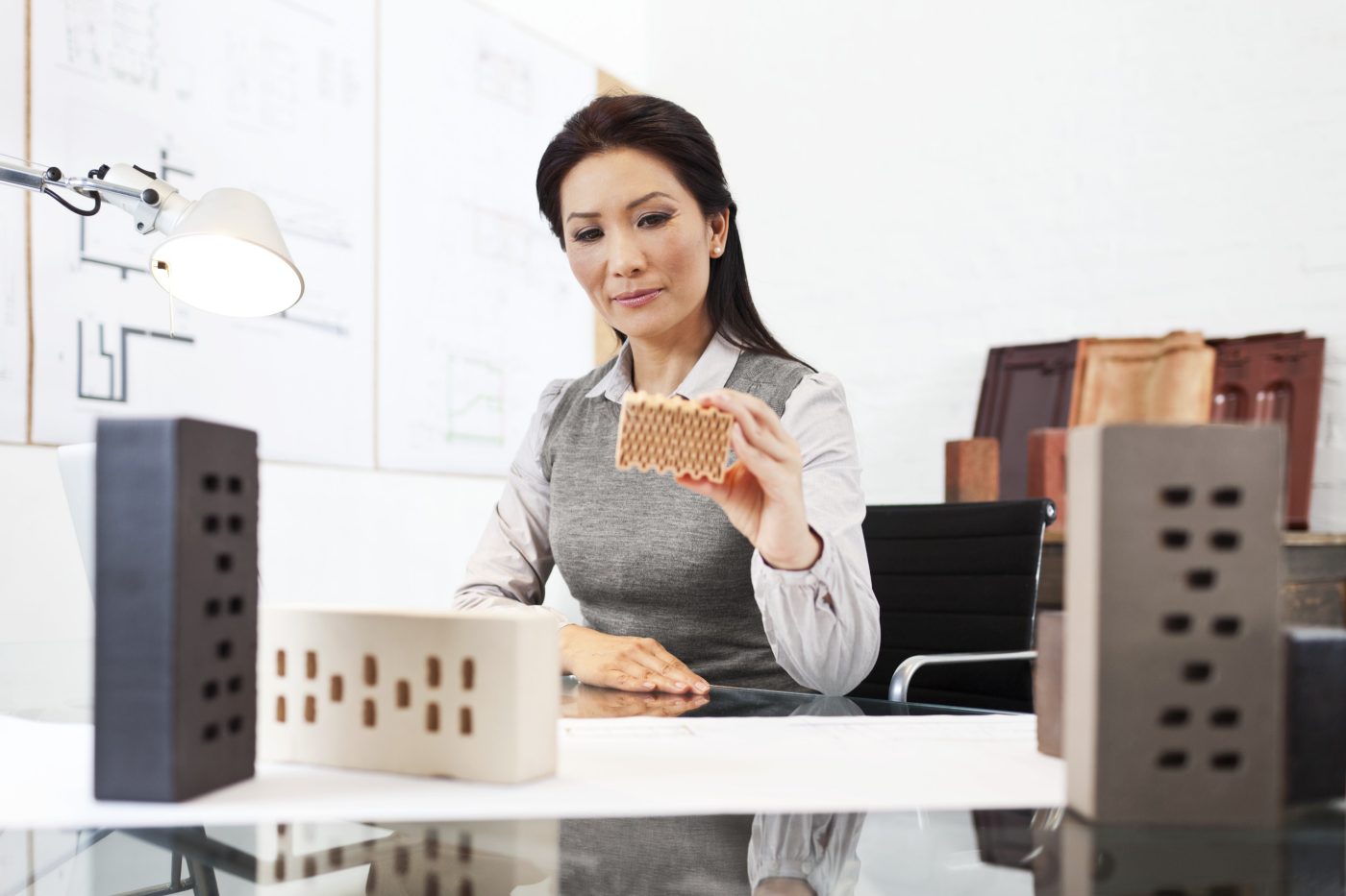 Female architect looking at miniature clay block surrounded by blueprints and bricks
