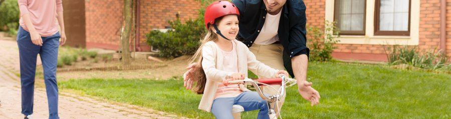 Father protecting little girl while learning to ride the bike while mother watches them