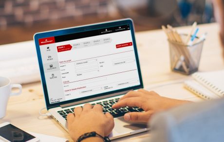 Young man working at modern office space, working on project. Sitting at the table and using laptop. Brick wall in background. Over the shoulder view.