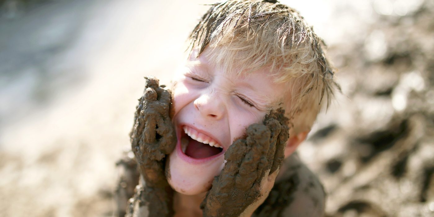 A cute little boy child is laughing as he plays outside in the mud and rubs dirt on his face with his hands.