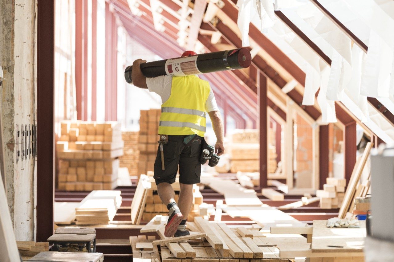 Urban roofer carrying underlay sheet on his shoulder in renovated attic wearing hard hat and safety jacket