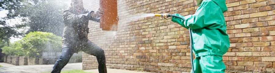 Boy hosing man holding a clay roof tile for protection in front of brick-built façade